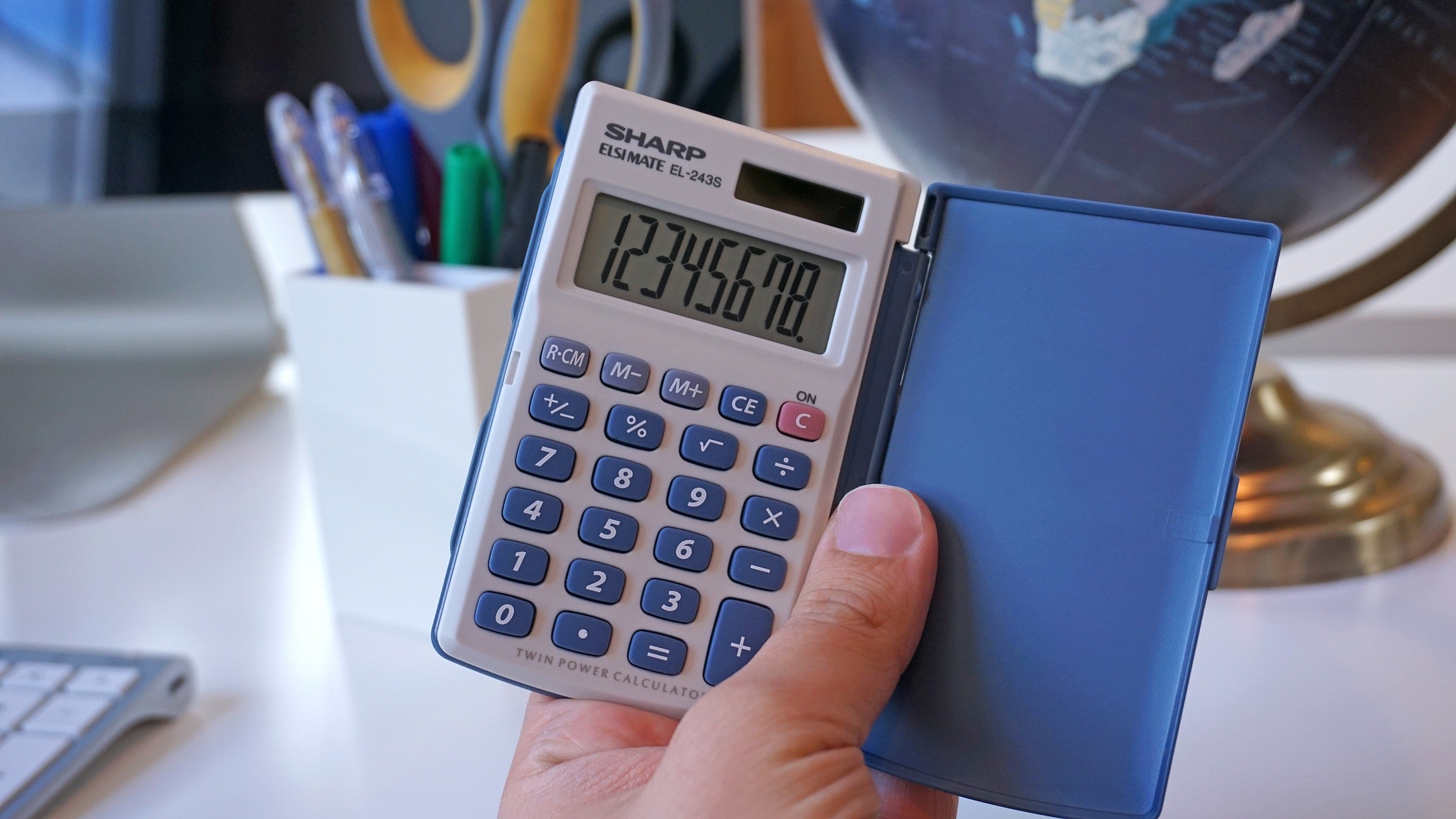hand holding a white and blue pocket calculator with a hinged case, globe and desktop supplies visible in the background